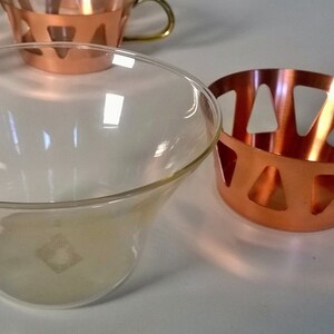 May include: A set of four clear glass cups with copper holders. The holders have a geometric design with cut-out triangles. The cups are perfect for serving cocktails or other beverages.