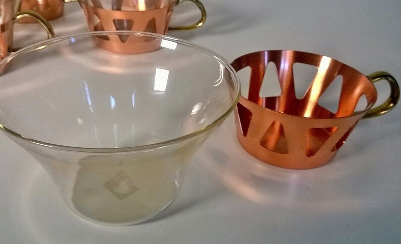 May include: A set of four clear glass cups with copper holders. The holders have a geometric design with cut-out triangles. The cups are perfect for serving cocktails or other beverages.