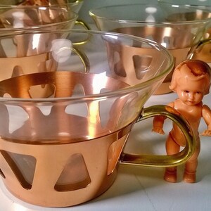 May include: Set of four copper and glass teacups with gold handles. The cups are arranged in a row, with a small doll standing in front of the cups.