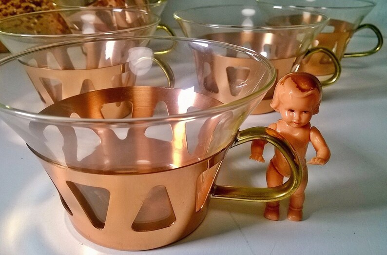 May include: Set of four copper and glass teacups with gold handles. The cups are arranged in a row, with a small doll standing in front of the cups.