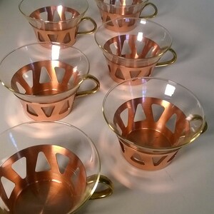 May include: Set of six clear glass cups with copper colored bands and handles. The cups have a geometric design.