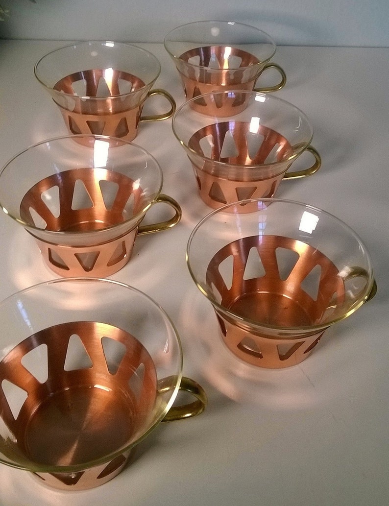 May include: Set of six clear glass cups with copper colored bands and handles. The cups have a geometric design.