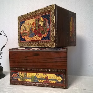 Peut inclure: Deux boîtes en étain vintage avec des motifs ornés. La boîte supérieure présente une peinture miniature persane colorée de personnages dans un jardin. La boîte inférieure a un design similaire avec le texte "150 Jahre Teehaus Ronnefeldt Frankfurt".