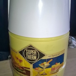 Könnte beinhalten: Eine gelbe und weiße isolierte Getränkebehälter mit einem Griff. Der Behälter hat ein Etikett mit der Aufschrift "Super Therm" und "Hält tagelang kalt". Das Etikett zeigt auch Bilder von Getränken und Desserts.