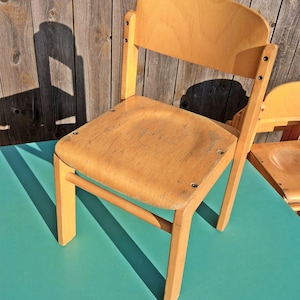 Op de afbeelding: Een kleine, lichtgekleurde houten stoel met een gebogen rugleuning en een afgeronde zitting. De stoel heeft vier rechte poten en een horizontale steunbalk. De stoel staat op een turquoise oppervlak, met een houten hek op de achtergrond.