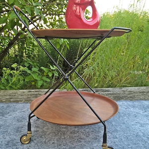 Puede incluir: Un carro de servicio de metal negro de dos niveles con bandejas de madera. El carro tiene cuatro ruedas doradas y está sobre una alfombra azul con estampado. Una jarra de cerámica roja con flores amarillas está en la bandeja superior.