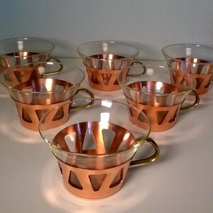 May include: Set of six clear glass cups with copper trim and brass handles. The cups have a geometric design with triangular cutouts.