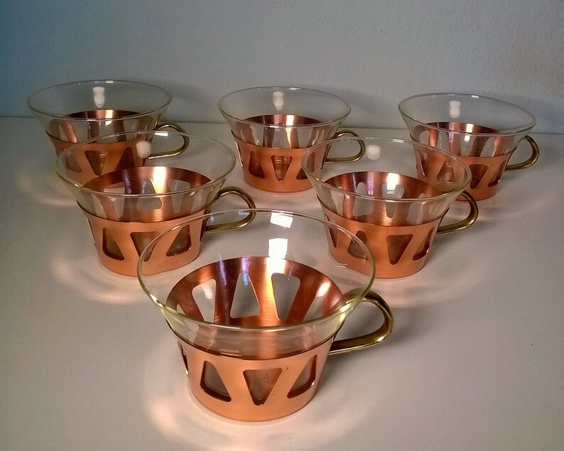 May include: Set of six clear glass cups with copper trim and brass handles. The cups have a geometric design with triangular cutouts.