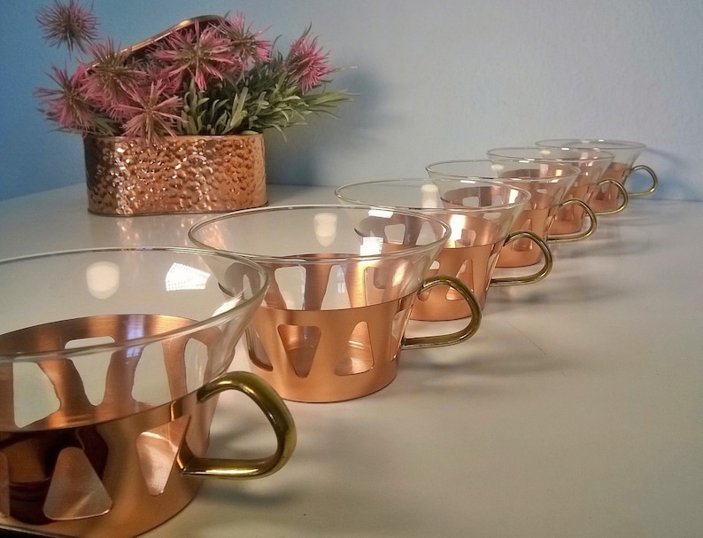 May include: Set of six copper and glass cups with brass handles. The cups have a unique design with cutouts in the copper band.
