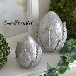 Könnte beinhalten: Zwei dekorative, silberfarbene Ornamente in Artischockenform. Die Ornamente sind aus einem strukturierten Material gefertigt und haben eine leicht abgenutzte Oberfläche. Die Ornamente stehen auf einem weißen Regal mit grünem Laub im Hintergrund.