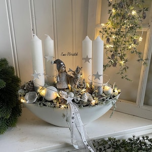 Könnte beinhalten: Weißes Keramik-Centerpiece mit vier weißen Kerzen, Sternakzenten und dekorativen Elementen. Das Centerpiece enthält eine kleine Figur, ein Rentier, Tannenzapfen und Lichterketten. Der Text "Ems-Floristik" ist sichtbar. Eine festliche Weihnachtsdekoration.
