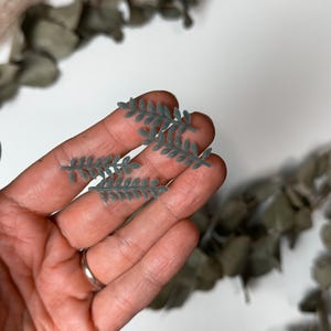 Könnte beinhalten: Nahaufnahme von kleinen, graugrünen, blattförmigen Verzierungen. Die filigranen, detaillierten Blätter werden in einer Hand gehalten, wobei ein silberner Ring sichtbar ist. Getrockneter Eukalyptus und anderes Grünzeug sind im Hintergrund zu sehen, was auf ein Bastel- oder Blumendesign hindeutet.