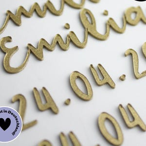 Könnte beinhalten: Goldfarbene, glitzernde, dreidimensionale Schreibschrift-Buchstaben, die den Namen Emilia und das Datum 24.04.20 bilden. Die Buchstaben haben eine texturierte Oberfläche und sind in Reihen angeordnet. Ein runder Aufkleber mit Herz ist zu sehen.