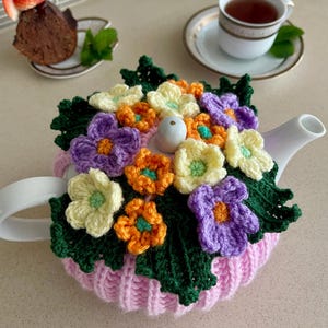 May include: A white ceramic teapot with a pink knitted cozy adorned with crocheted flowers in shades of purple, orange, and yellow. A cup of tea and a small cake with a strawberry garnish are also visible.