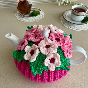 May include: A white ceramic teapot adorned with a vibrant, handmade crochet cozy. The cozy features a bright pink base, green leaves, and delicate pink and white flowers. A charming addition to any tea set.