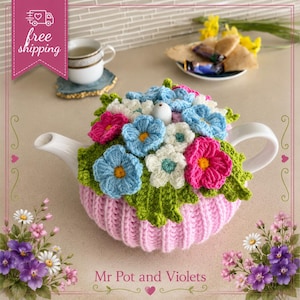 May include: A white ceramic teapot covered by a crocheted cozy featuring a ribbed light pink base and a top layer of textured green leaves adorned with vibrant blue, white, and magenta crocheted flowers with yellow and teal centers.