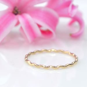 Könnte beinhalten: Ein dünner goldener Ring mit einem wellenförmigen, strukturierten Design. Der Ring liegt auf einer weißen Oberfläche.