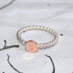 Könnte beinhalten: Ein silberner Perlenring mit einer roségoldenen Scheibe mit dem Buchstaben "S". Der Ring ist vor einem weißen Hintergrund mit schwarzen Tintenpinselstrichen platziert. Ein zarter Schmuck.