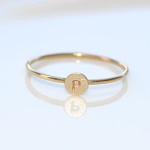 Könnte beinhalten: Ein zarter goldener Ring mit einem kleinen, kreisförmigen Anhänger mit dem eingravierten Buchstaben "P". Der Ring hat ein schmales Band und eine polierte Oberfläche, die das Licht reflektiert. Der Ring ist ein personalisiertes Schmuckstück.