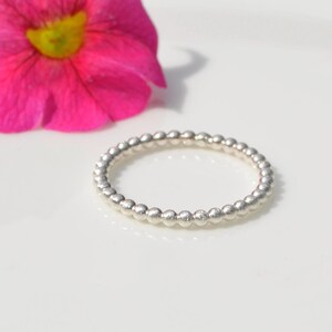 Könnte beinhalten: Ein Silberring bestehend aus einem durchgehenden Band kleiner, gleichmäßiger, kugelförmiger Perlen in kreisförmiger Anordnung. Der Ring liegt neben einer leuchtend pinkfarbenen Blume mit gelber Mitte, die die metallische Textur betont.