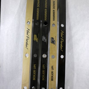 May include: Four wooden game pieces with black and gold paint. The pieces have holes drilled into them and the text "Hail Purdue!" and "Boiler Up!" is printed on them. The text "Hammer Down!" is printed on the top of each piece.