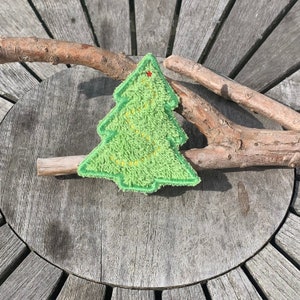 Può includere: Una toppa a forma di albero di Natale verde e soffice con dettagli ricamati gialli e rossi. La toppa è su una superficie di legno invecchiato con rami d'albero.