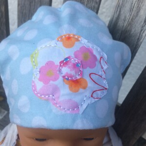 Op de afbeelding: Een lichtblauwe baby muts met witte stippen en een bloemenapplicatie. De bloem is gemaakt van witte stof met roze, oranje en rode bloemen en een wit centrum.