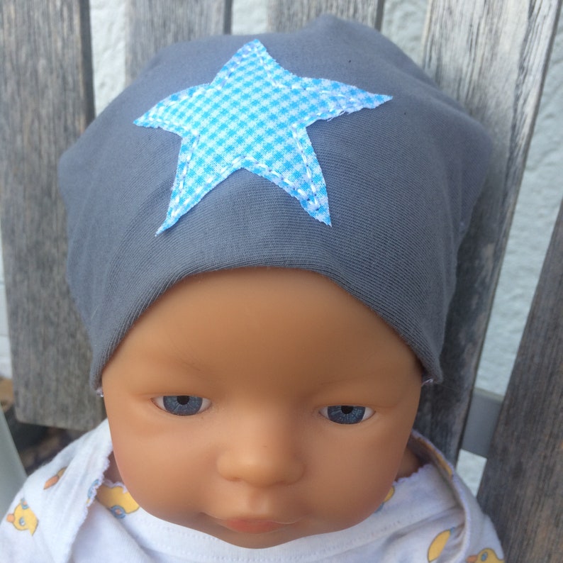 Op de afbeelding: Een grijze baby muts met een blauwe en witte geruite ster aan de voorkant.