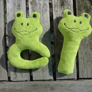 May include: Two green plush frog toys. One frog is shaped like a ring and the other is a long, thin toy. Both frogs have black eyes and a stitched-on smile.