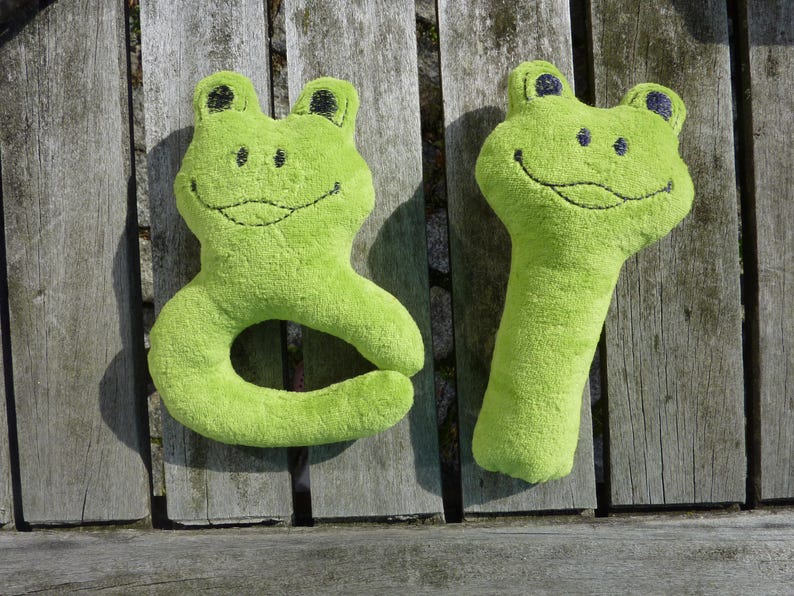 May include: Two green plush frog toys. One frog is shaped like a ring and the other is a long, thin toy. Both frogs have black eyes and a stitched-on smile.
