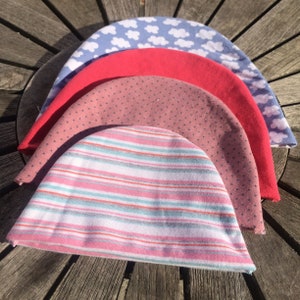 May include: Four knitted baby hats stacked on top of each other. The top hat is light blue with a white cloud pattern. The second hat is red. The third hat is pink with white polka dots. The bottom hat is white with pink and blue stripes.