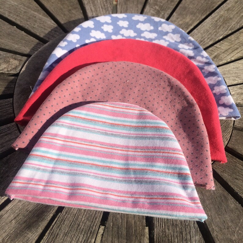 May include: Four knitted baby hats stacked on top of each other. The top hat is light blue with a white cloud pattern. The second hat is red. The third hat is pink with white polka dots. The bottom hat is white with pink and blue stripes.