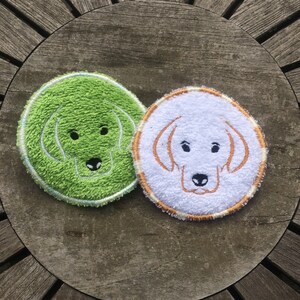 May include: Two round, white, terry cloth coasters with embroidered dog faces. One coaster is green with a white outline and the other is white with a gold outline.