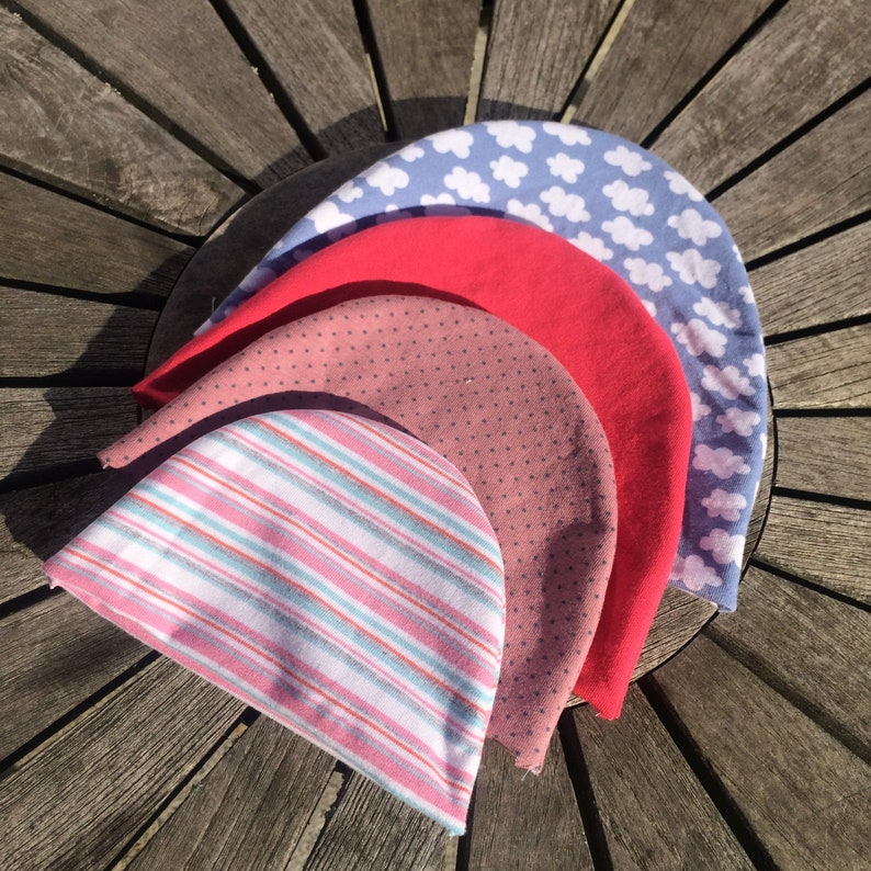 May include: Five fabric hats stacked on top of each other. The top hat is blue with white clouds, the second hat is red, the third hat is pink with white polka dots, the fourth hat is pink and white striped, and the bottom hat is pink with white polka dots.