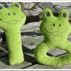 May include: Two green plush frog toys. One is a rattle with a long body and the other is a ring toy with a round body. Both have embroidered eyes and a smiling mouth.