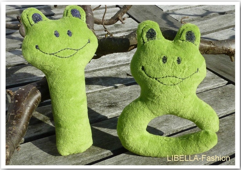 May include: Two green plush frog toys. One is a rattle with a long body and the other is a ring toy with a round body. Both have embroidered eyes and a smiling mouth.