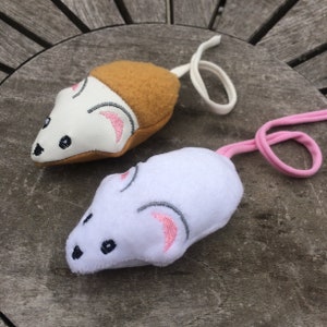 May include: Two plush cat toys shaped like mice. One mouse is white with brown fur on its back and a white string. The other mouse is white with a pink string.