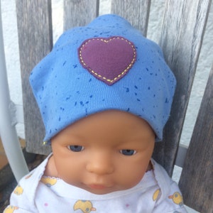 Op de afbeelding: Een blauwe baby muts met een paars hartje erop gestikt. De muts heeft een gespikkeld patroon.