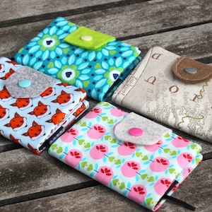 May include: Four small fabric-covered notebooks with different patterns. The notebooks have a flap with a button closure. The patterns include foxes, flowers, a map, and a geometric design. The notebooks are on a wooden surface.