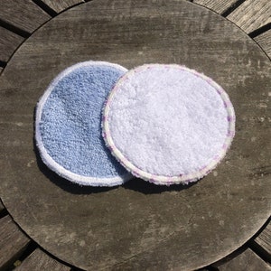 Puede incluir: Dos almohadillas redondas reutilizables para desmaquillar. Una es azul claro con costuras blancas y la otra es blanca con costuras moradas.