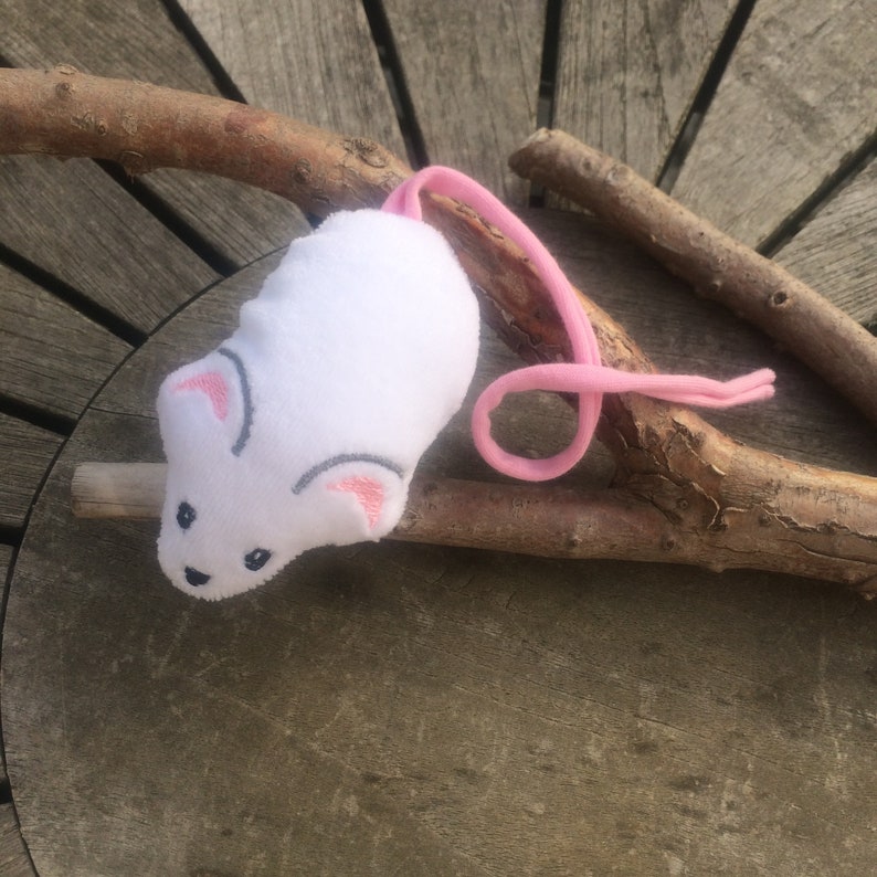 May include: White plush toy mouse with pink ears and a pink tail. The mouse is lying on a wooden branch.
