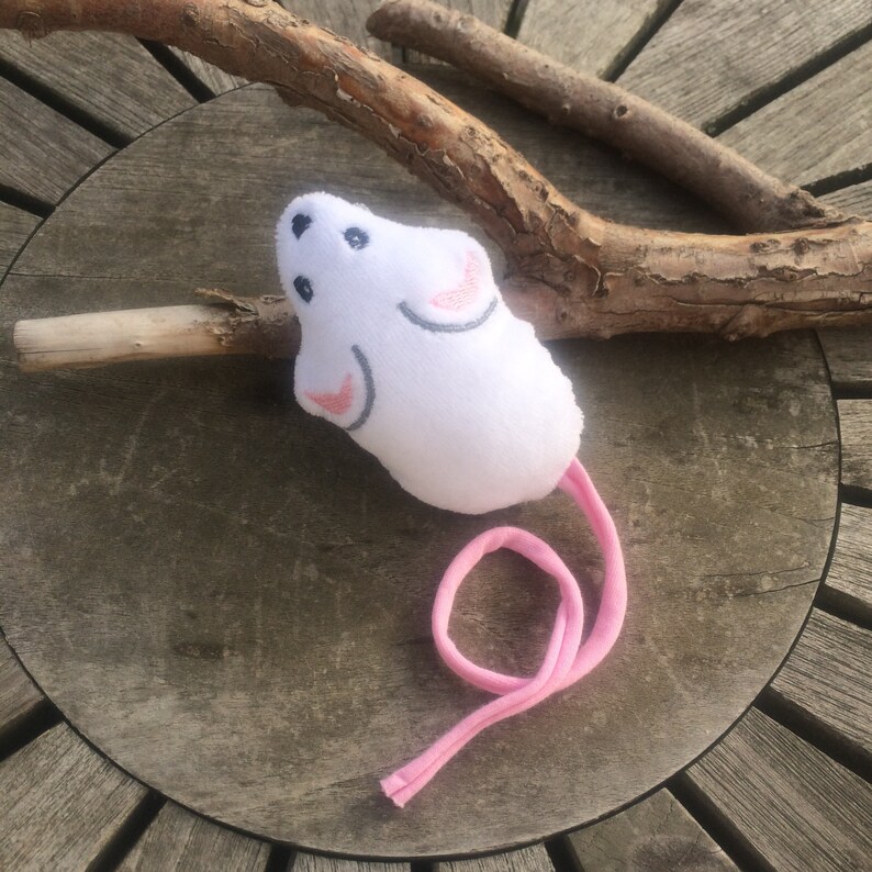 May include: White plush toy mouse with pink ears and a pink tail. The mouse is lying on a wooden surface.