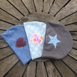 Op de afbeelding: Drie stoffen hoofdbanden met verschillende ontwerpen. Een hoofdband is blauw met witte stippen en een roze bloemenapplicatie. Een andere hoofdband is blauw met witte vlekken en een bruine hartapplicatie. De derde hoofdband is grijs met een blauw-wit geruite sterapplicatie.