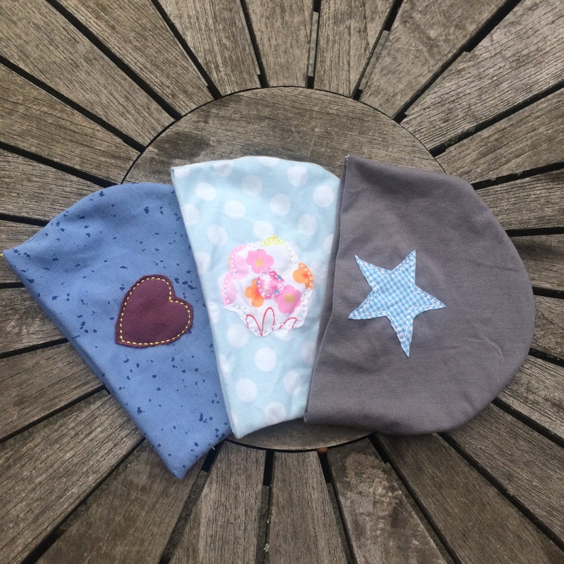 Op de afbeelding: Drie stoffen hoofdbanden met verschillende ontwerpen. Een hoofdband is blauw met witte stippen en een roze bloemenapplicatie. Een andere hoofdband is blauw met witte vlekken en een bruine hartapplicatie. De derde hoofdband is grijs met een blauw-wit geruite sterapplicatie.