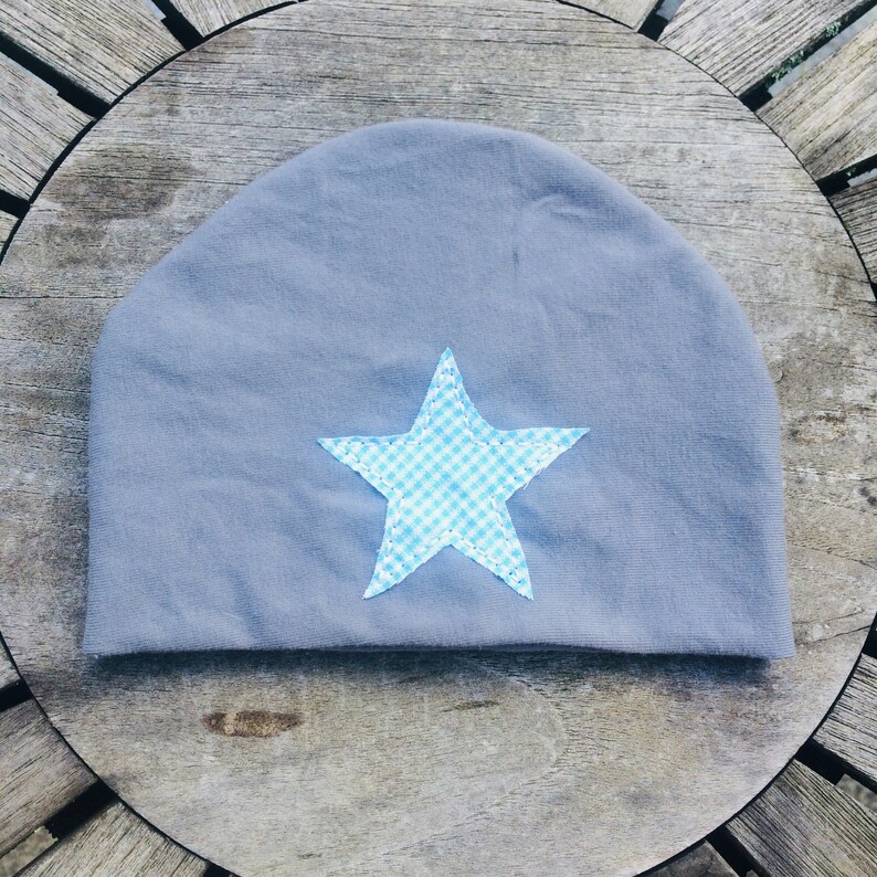 Op de afbeelding: Grijze beanie met een blauwe en witte geruite ster die op de voorkant is genaaid.