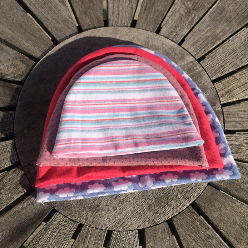 May include: Four colourful knitted hats stacked on top of each other. The top hat is pink and white striped, the second hat is brown with white polka dots, the third hat is red, and the bottom hat is blue with white clouds.
