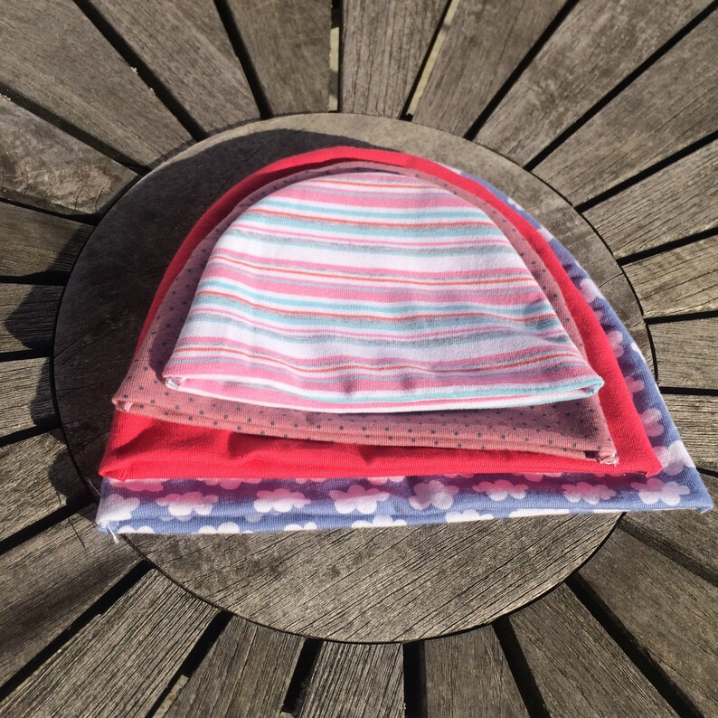 May include: Four colourful knitted hats stacked on top of each other. The top hat is pink and white striped, the second hat is brown with white polka dots, the third hat is red, and the bottom hat is blue with white clouds.