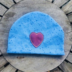 Op de afbeelding: Een blauwe beanie met een klein, hartvormig plekje dat op de voorkant is genaaid. Het plekje is donker bordeauxrood met gele stiksels rond de randen.