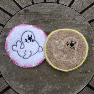 May include: Two round, white and brown fabric coasters with embroidered seal designs. The white coaster has a pink border and the brown coaster has a yellow border.