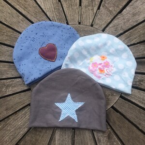 Op de afbeelding: Drie verschillende stoffen mutsen met verschillende versieringen. E&eacute;n muts is blauw met witte stippen en een bruin hart, een andere is lichtblauw met witte stippen en een roze bloem en de laatste is grijs met een blauwe ster.